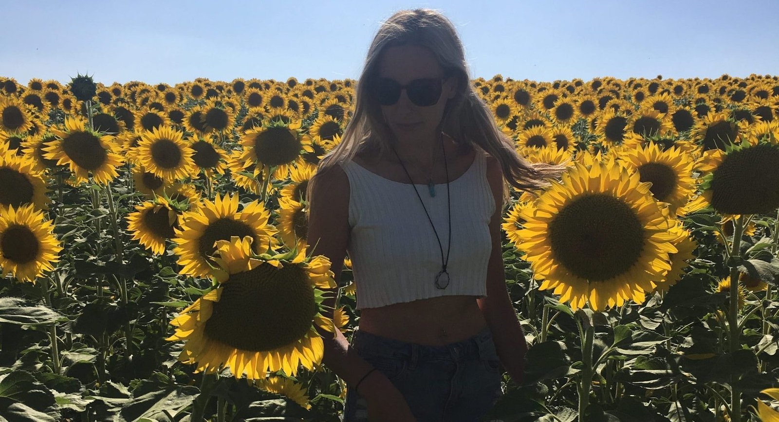 blonder girl wearing sun glasses and a white crop top in a field of yellow sunflowers